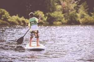 Child standing on a surfboard floating on a river paddling the water.