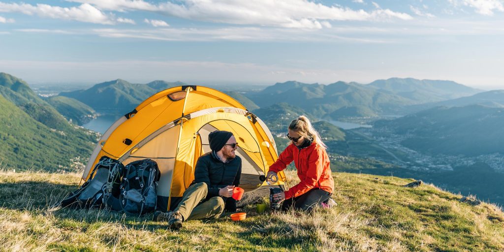 Man and woman couple siting in front of their yellow pitched tent overlooking a mountainous valley.
