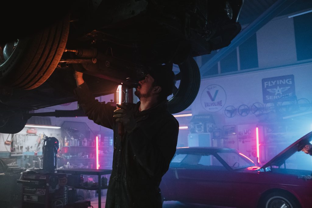 Chevrolet vehicle lifted up allowing a Chevrolet certified mechanic to walk under to do a full vehicle inspection. Scene is a Smokey mechanic shop with pink and orange neon lights in the background.