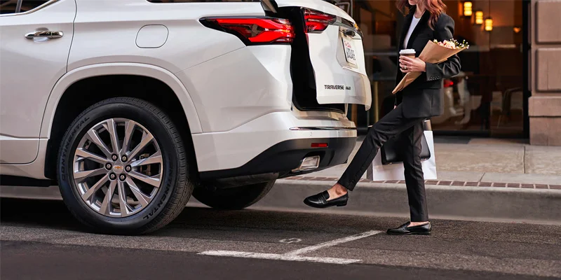 photo of the back half of a brand new white 2023 Chevrolet Traverse. A woman has her hands full walking up to her brand new Traverse and is able to use her foot to tap the bottom of the bumper, opening up the back door to set her stuff down.